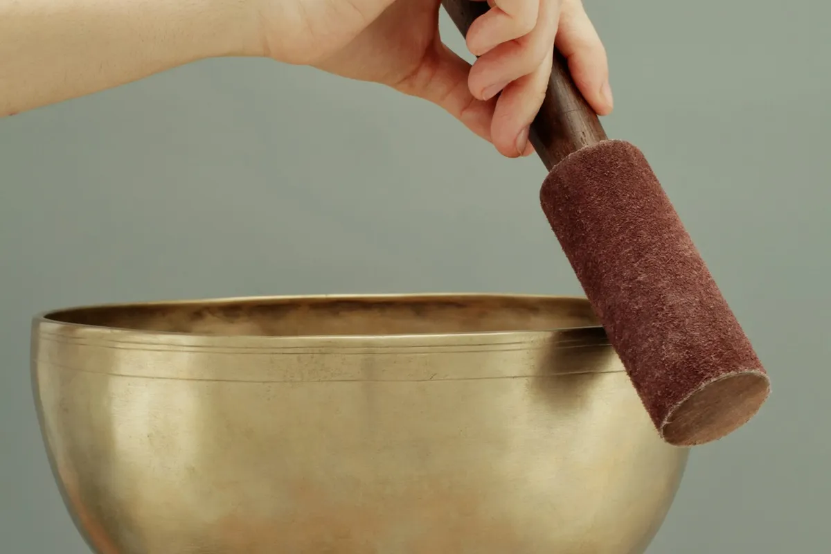 Hand-hammered singing bowl held in two hands with a felt-covered mallet against a calm sage background