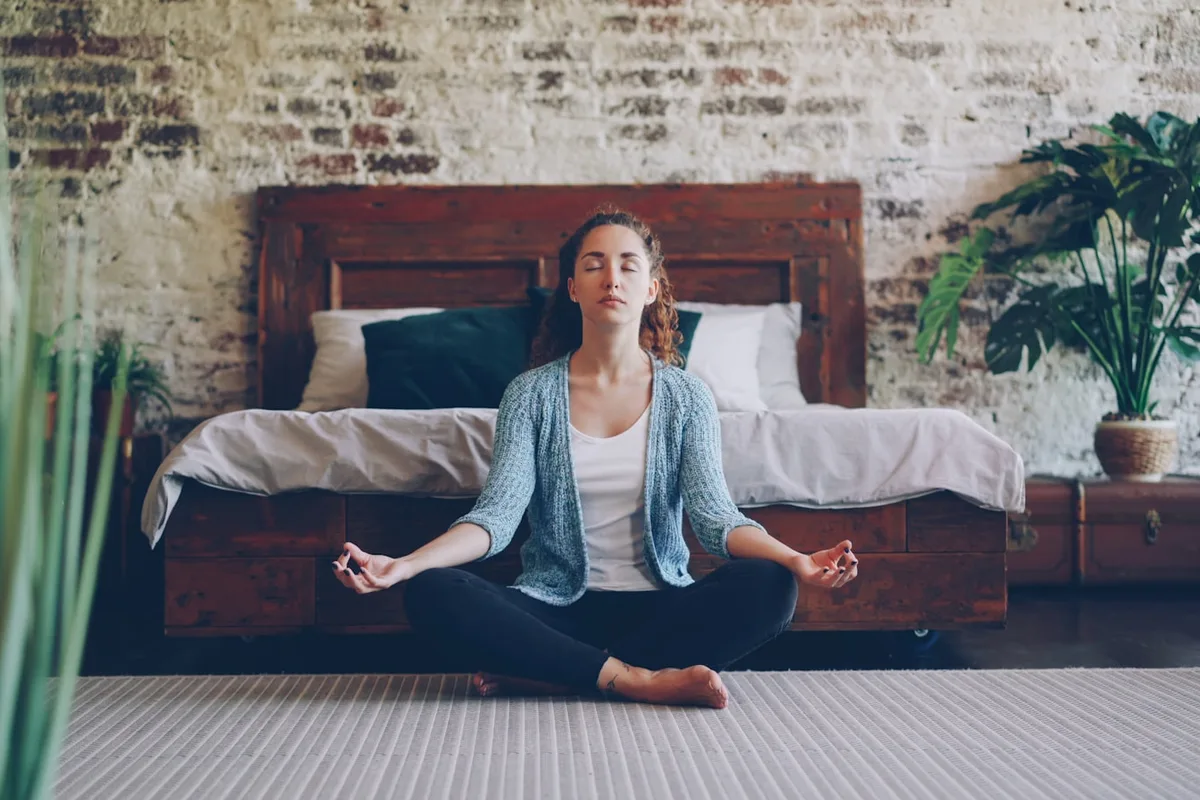 Woman meditating cross-legged with eyes closed in a warm, plant-filled room