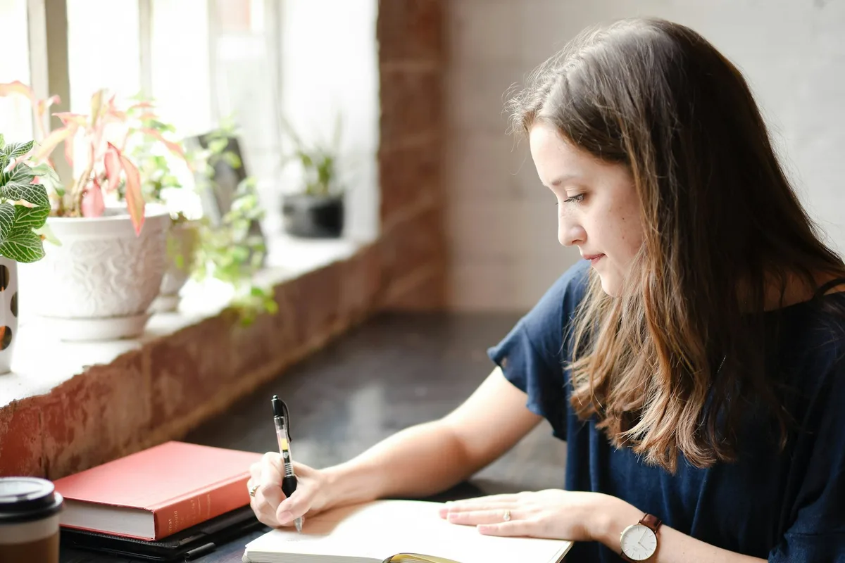 Woman journaling in a notebook by a plant-filled window in warm light